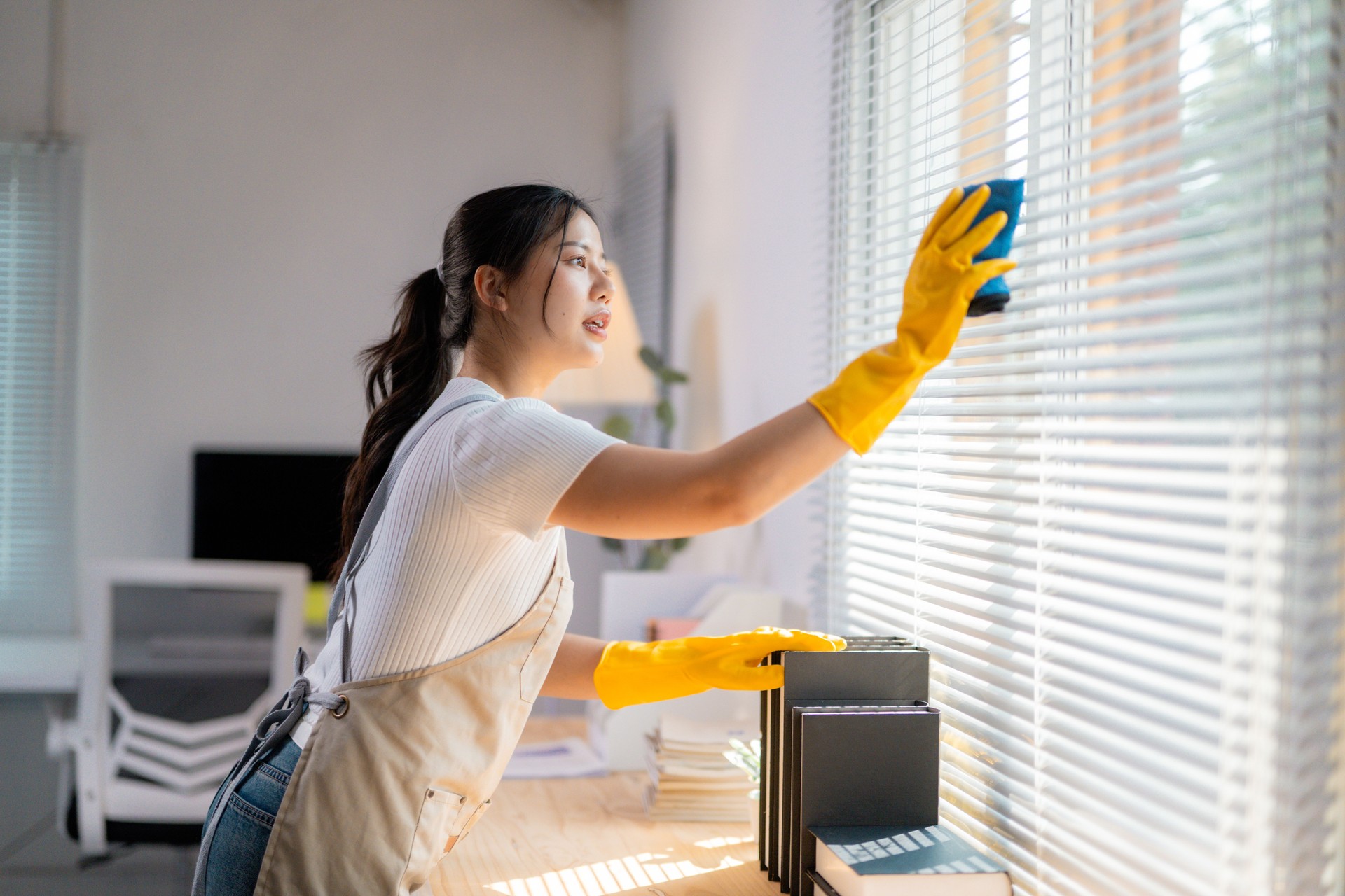 Professional Asian housekeeper wiping office window blinds, wearing protective gloves and apron, ensuring spotless workplace cleanliness